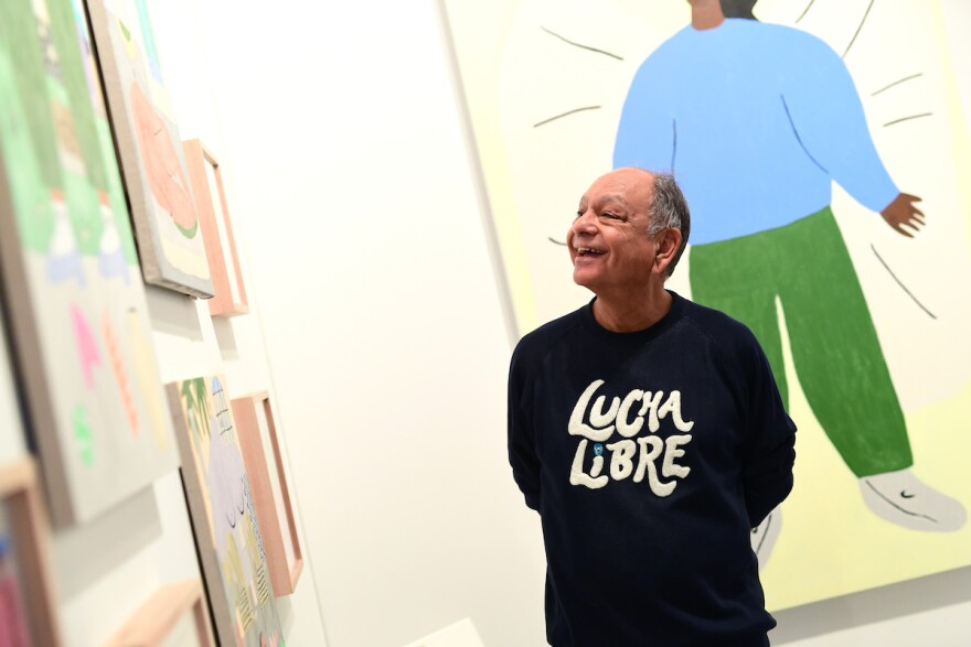 Cheech Marin in a black t-shirt that reads "Lucha Libre" stares at a wall of art.