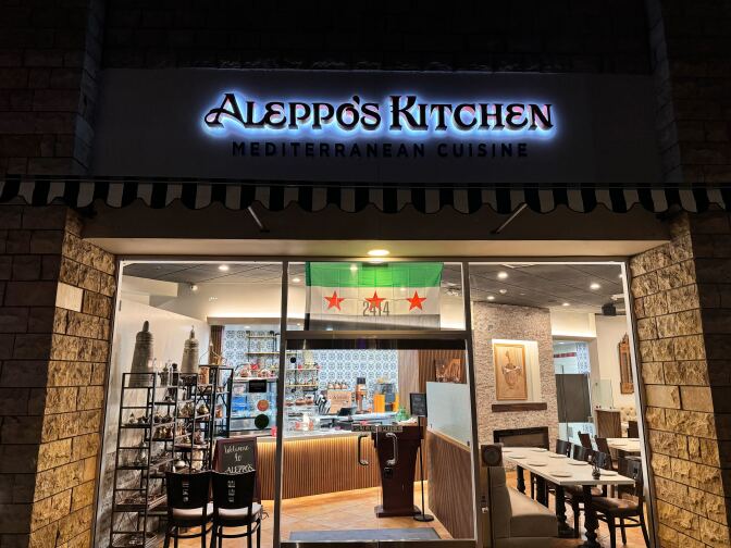 A black, green and white flag hangs outside a restaurant. The words Aleppo's Kitchen are backlit on the restaurant sign.