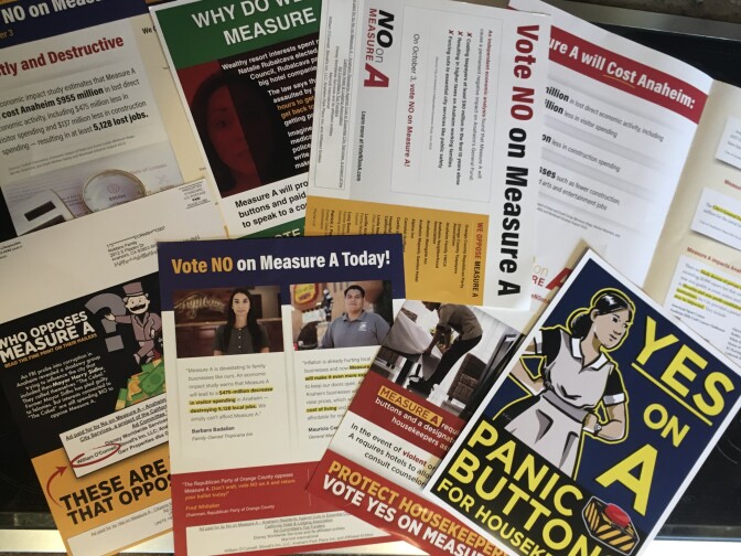 Campaign mailers spread across a table. One has a drawing of a housekeeper and says "Yes on A. Panic Button for Housekeepers." Another says "Vote NO on Measure A Today!"