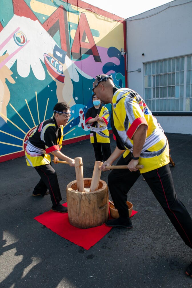 Two Kodama Taiko performers in yellow and black happi coats crouch over a wooden mortar, pounding mochi with wooden mallets in synchronized motion while a third performer observes, all set against a colorful street mural.
