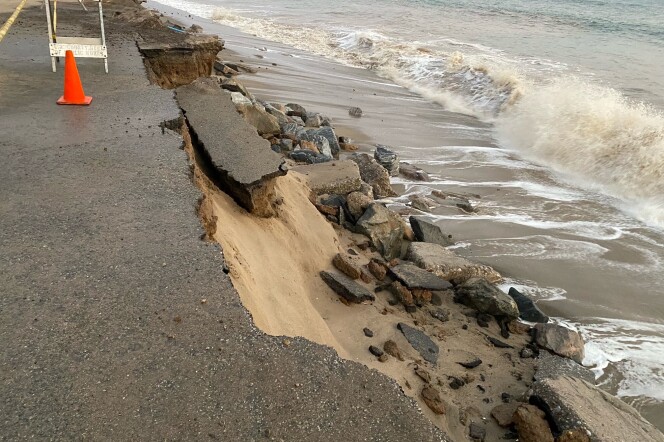 The side of the roadway has shattered and dropped into the sand as waves roll to shore. A caution cone and hazard sign are in the roadway.