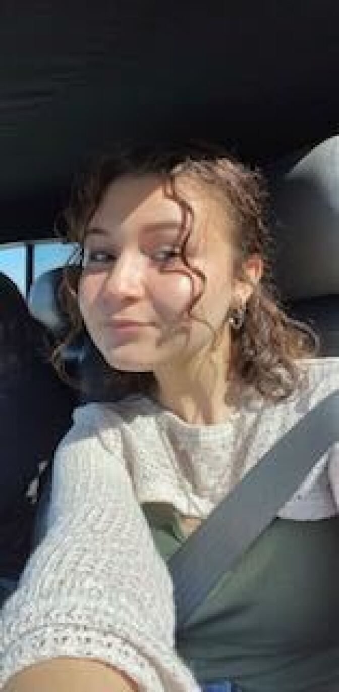 A young woman with curly hair posing with a half smile in a car with a seatbelt across her chest. 