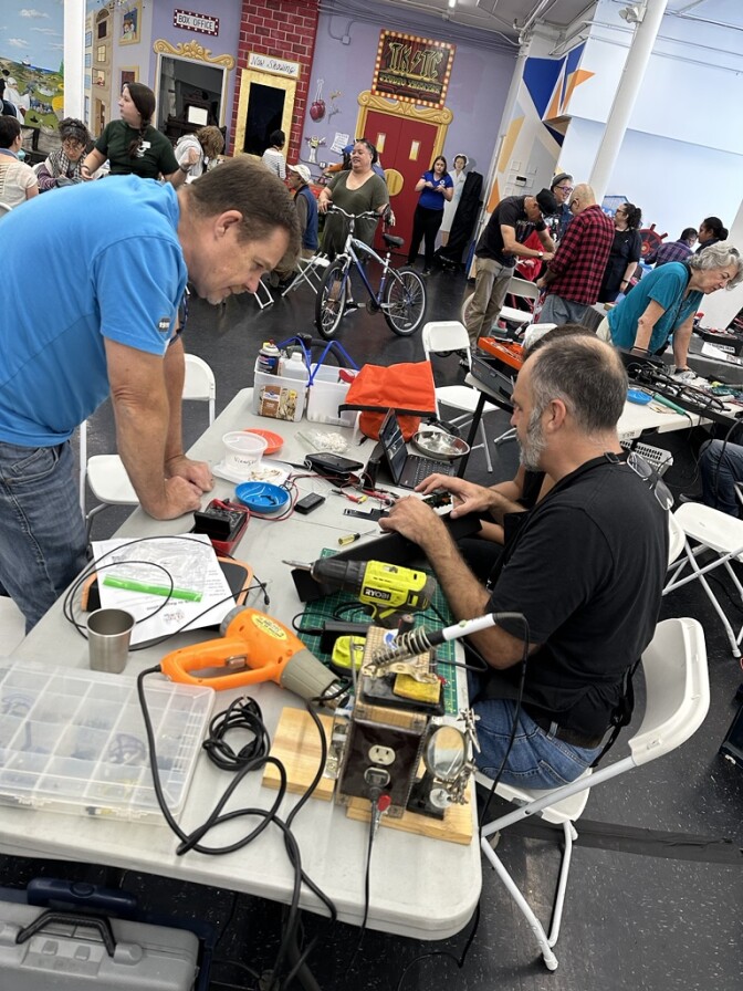 A room full of men and women of all ages. A white foldable table is set up closer to the camera, with two people seated behind it, including a man in blue jeans and a black t-shirt. Another man wearing a blue t-shirt and jeans is leaning over the table, which is covered in tools and electrical equipment.