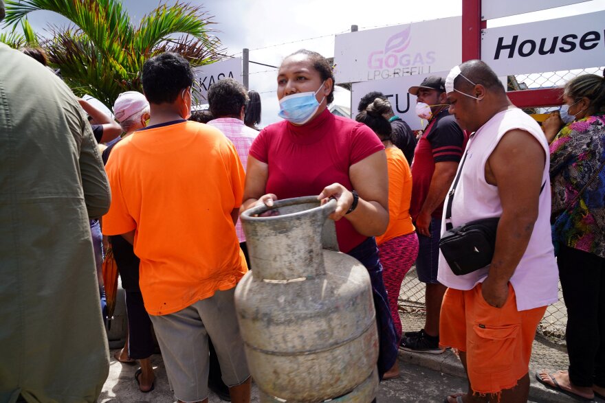 A woman in a red blouse with a medical mask hanging below her nose carries a metal gas container. She is surrounded by many other people.
