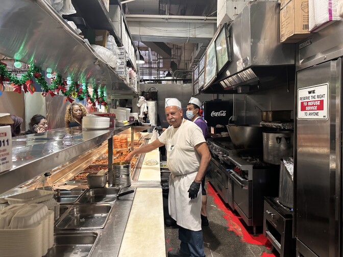 A man wearing a cook's cap and a white apron, and a black disposable glove stands behind the well-worn counter of a food stall known for it's Mexican food. Customers are waiting in line to order, while others are looking at tray after tray of Mexican food offerings, from pork to tongue and rice and more. A second man, wearing a mask over his nose and mouth, is also tending to the counter. 