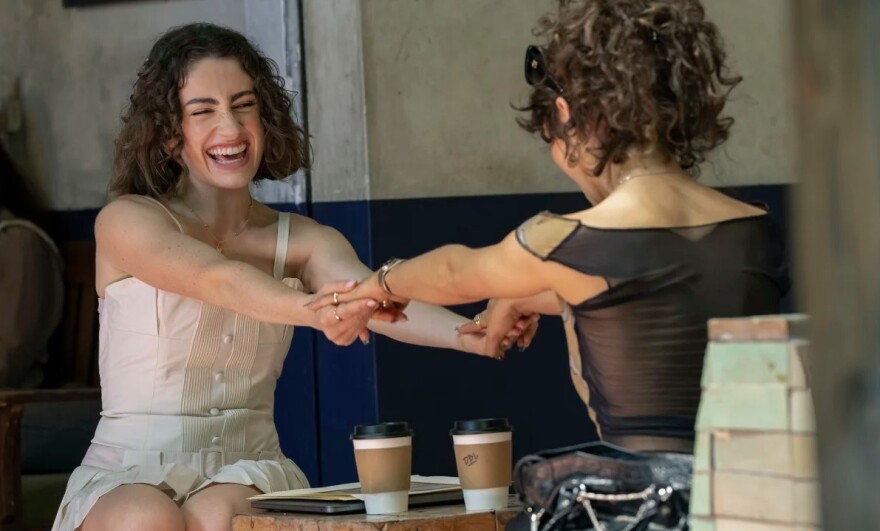 Two women laugh and hold hands while sitting outside in a booth. There are two coffees between them