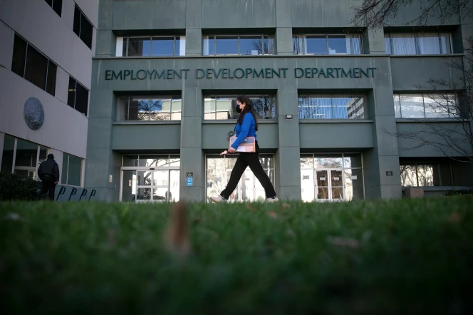 A woman wearing a blue long sleeved top and black pants walks past a large, dark green building with signage that reads, "Employment Development Department"