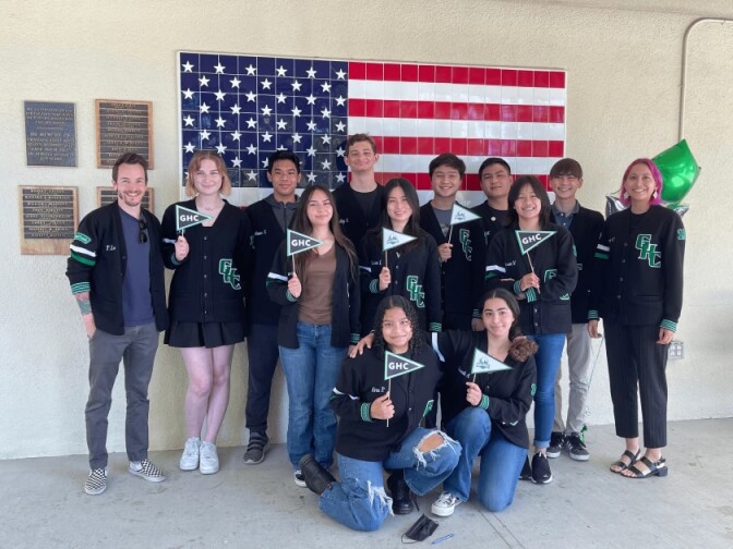 A group of Academic Decathlon students hold flags while posing for a photo in front of an American Flag.