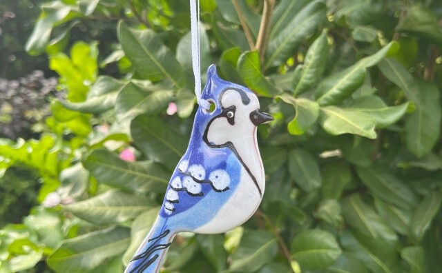 A hand crafted ceramic ornament depicting a blue jay hangs in a tree. 