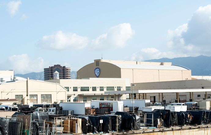 A wide view of several large beige warehouse-style buildings, one with the Warner Bros. logo. In front of the buildings are large trucks and movie equipment, mostly draped with black cloths. 