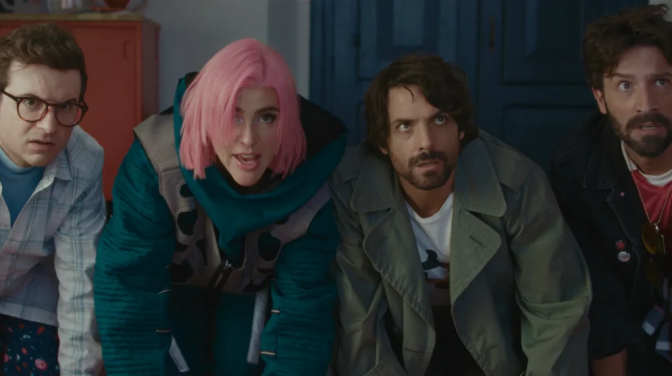 Four people lean forward with startled expressions, one wearing a pink wig and teal jacket, in a dimly lit room.