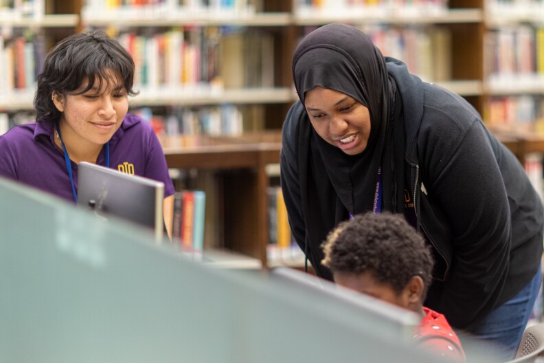 A Black feminine presenting person wearing a black hijab and black sweater bends over towards a Black child who is at a computer screen. On the left of frame a feminine presenting person with medium skin tone wearing a purple shirt and short dark hair looks on at the child and smiles.