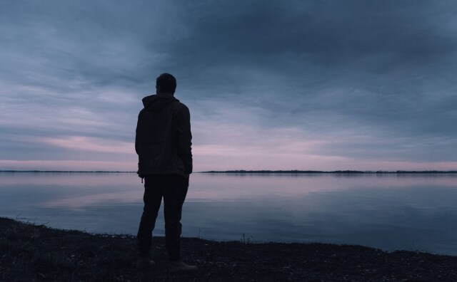 A silhouette of a man, wearing what appears to be a jack, at dusk looking out over a body of water