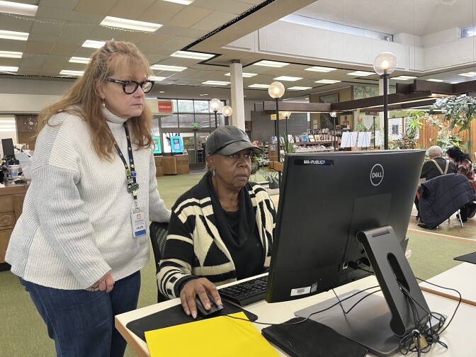 A white middle-aged woman helps a Black middle-aged woman at a desktop computer.