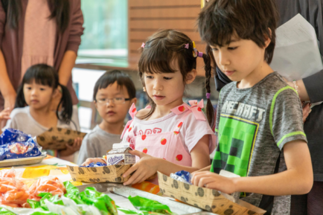 Four children pick up lunches from a line. One of the children wears a grey and green Minecraft tshirt and has short black hair. Another child has their in braids and wears pink overalls.  
