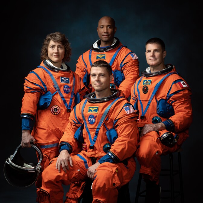 Four people are pictured in orange space suits: from left, a woman with light-tone skin, a man with dark-tone skin, a man with light-tone skin and a man with light-tone skin. The woman holds a helmet in her right hand.