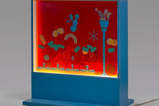 A standing, blue box holding illuminated red, yellow, green and blue artwork. It's displayed in a gray room. 
