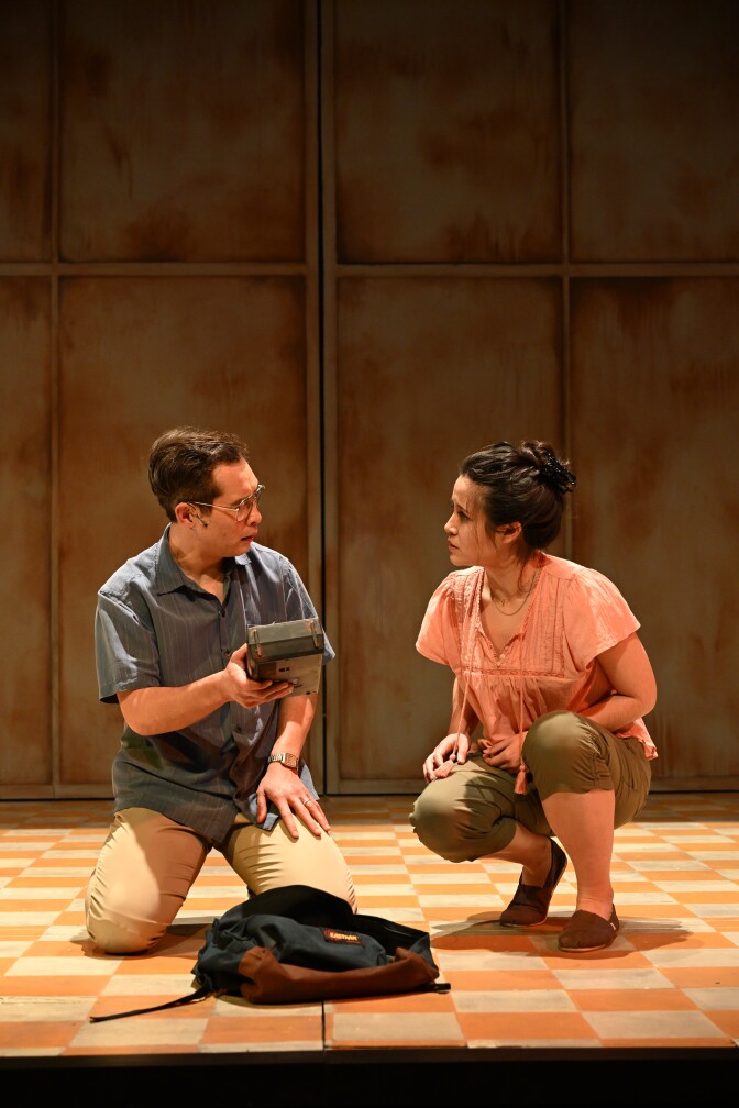 A man and woman in casual shirts and pants crouch on a stage with a checkerboard floor looking at each other. 