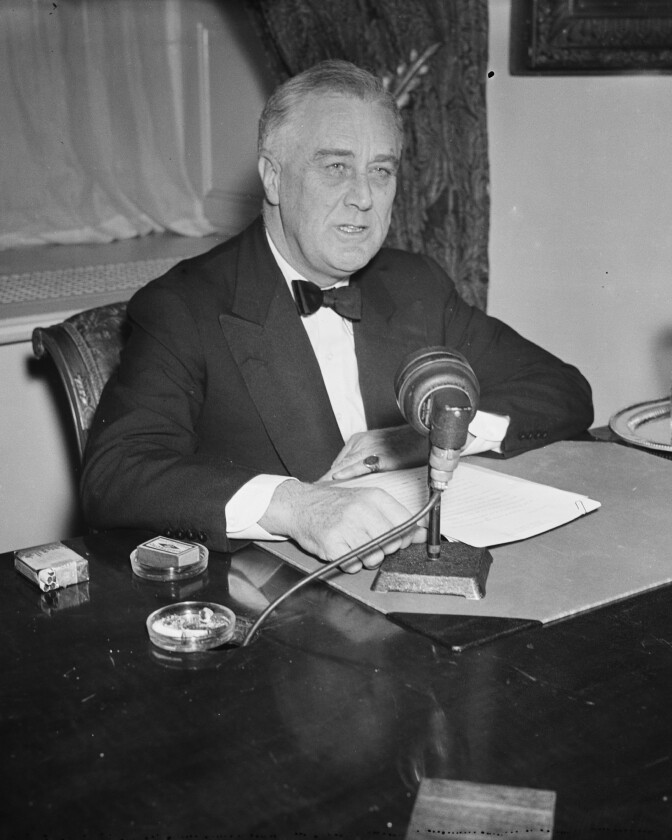 Black and white photo of a man wearing a tuxedo, sitting at a desk with a microphone on it