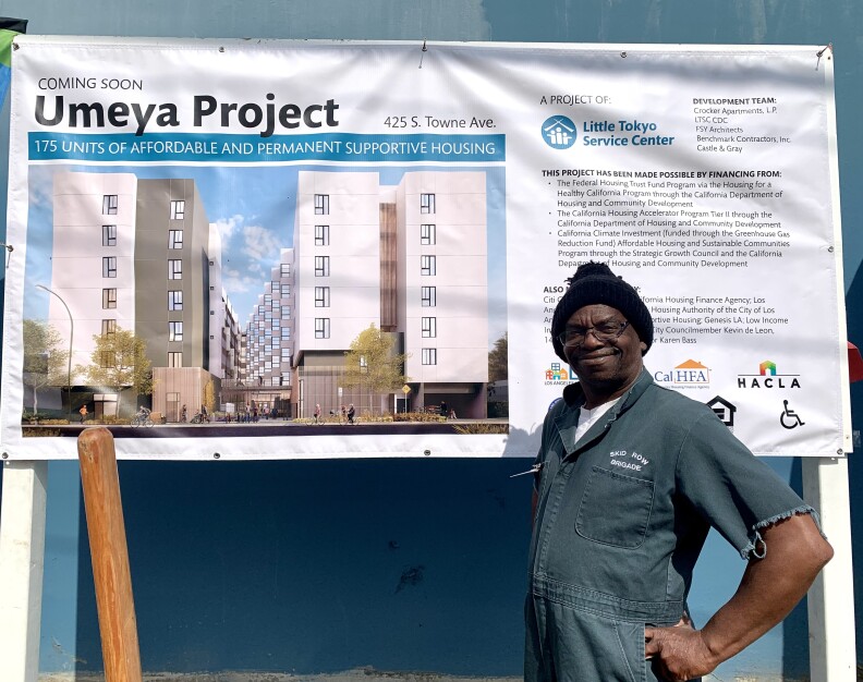 A Black man wearing glasses stands in front of a poster featuring an artist rendering of a white-walled housing complex.
