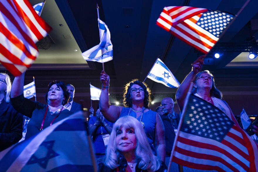 People wave Israeli and U.S. flags. Some are sitting and others standing. They appear to be lighted from a stage in front of them.