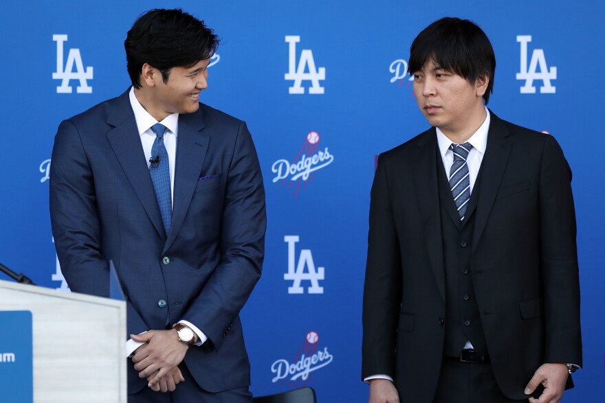 Two men are standing in front of a deep blue wall with the Los Angeles Dodgers logo plastered around. The man on the left is slightly taller and wearing a navy blue suit as he smiles and looks towards his right. The man on the right is wearing a black suit with a striped blue and white tie and has a neutral expression on his face.