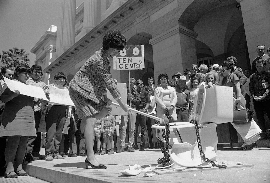 A black and white photo of a Chinese American woman in a skirt suit bending down with a hammer to smash a chained-up toilet. A group of people are around her with protest signs saying ten cents by the capitol building.