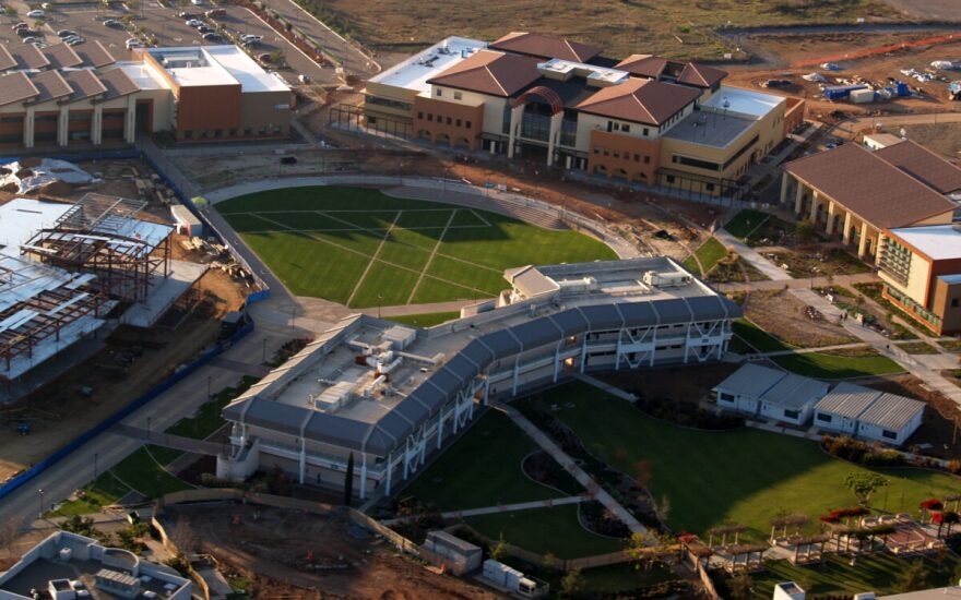 An aerial view of a university campus with a baseball diamond at the center. Several very large buildings encircle the diamond, and a parking lot can be seen in the upper left-hand corner. It is day time.