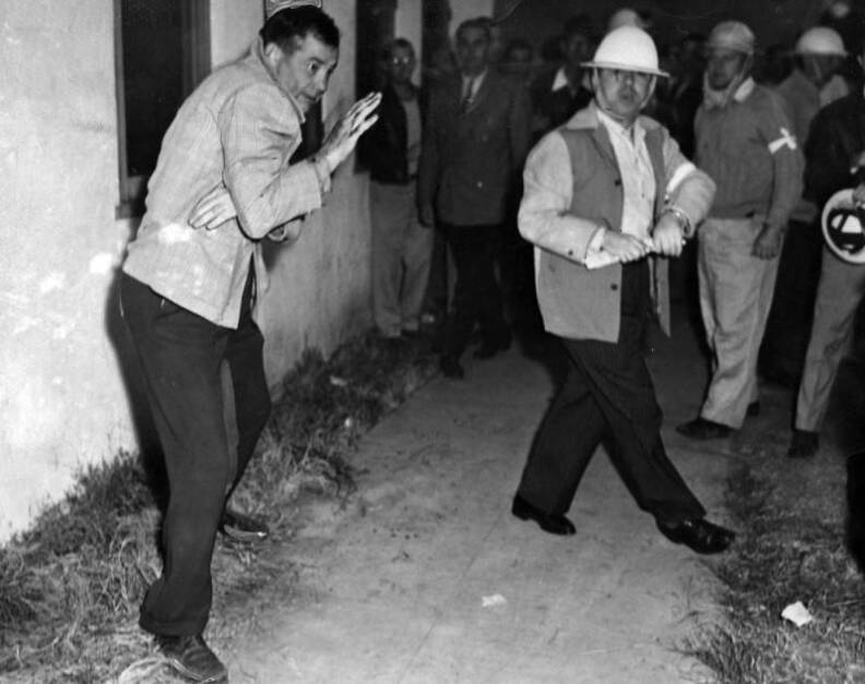 The man is shown in front of a building. Several other men are in the background, some with white hard hats.