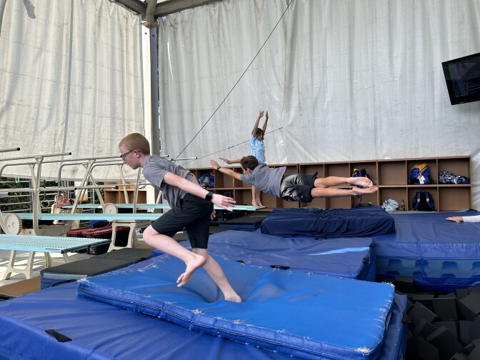 Blue mats are piled on the floor with three diving boards along the left side. In the foreground, a boy with light skin and glasses looks like he's been caught in motion, running on top of one the mats. Another boy is caught in mid-air parallel to the mats, and a third child, in the background, is standing on the diving board on their tippy toes, arms pointed to the sky. 