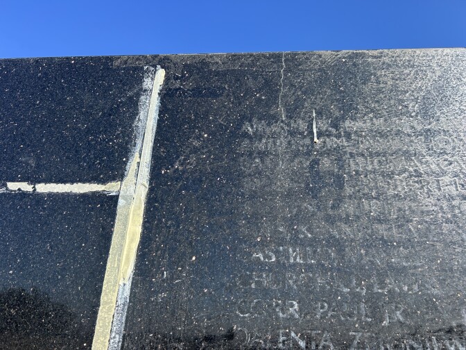 A closeup of a hairline crack running through black granite. The outline of names are visible on the granite and there is leftover masking tape next to the names.