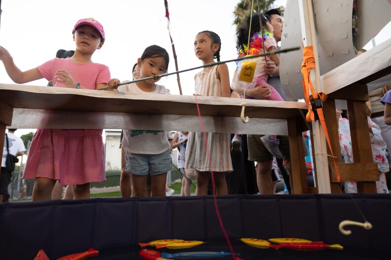 Three girls with fishing poles lean over a large tub with floating wooden red and yellow fish.