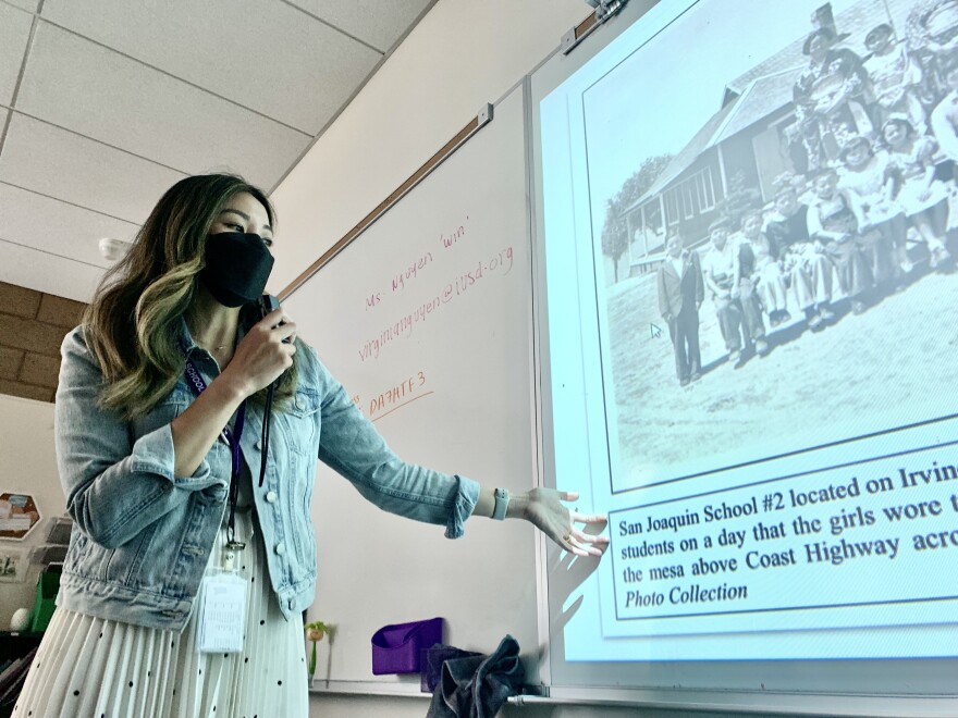 An Asian American woman with ombre dyed hair and wearing a jean jacket stands before a whiteboard onto which a black-and-white photo of Japanese American schoolchildren has been projected.