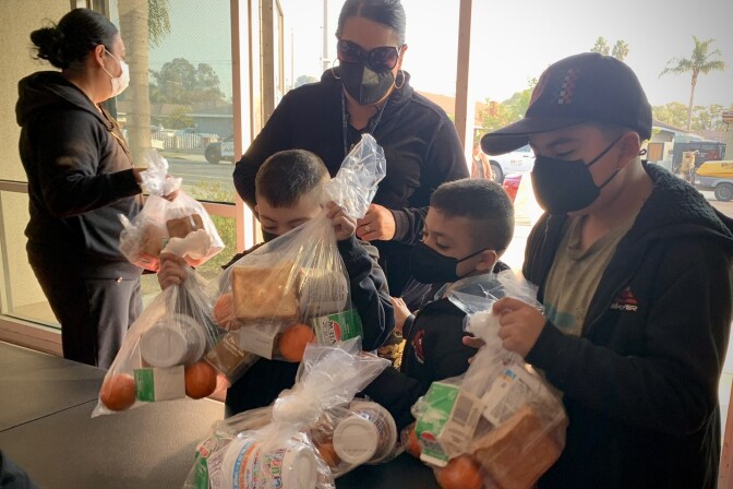 A woman and three boys with medium light skin tone wear black protective face masks and pick up six clear plastic bags containing oranges, milk, cereal and sandwiches. A woman with medium light skin tone, hair in a bun carries more bags in in the background.
