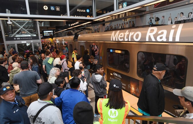 A subway train with the words "Metro Rail" on one of the carts arrive at the station. Many people are on the platform waiting.