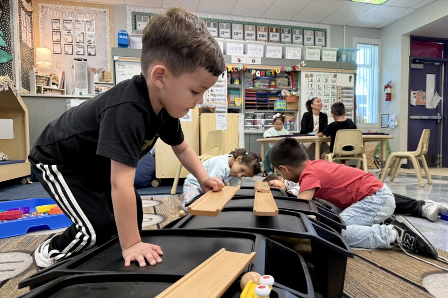  A student with light skin tone and black track pants and t-shirt picks up a wooden track. In the background, students with a variety of skin tones look at other parts of the track. 