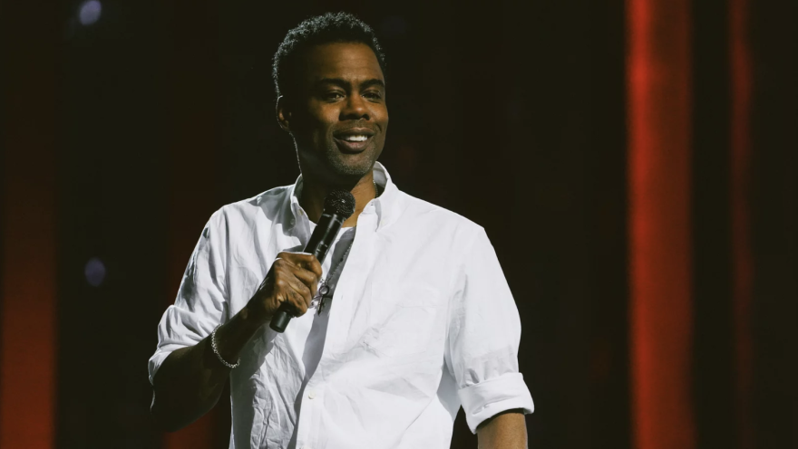 A man was dark skin tone is wearing a white shirt while speaking into a microphone he's holding in his right hand on stage.