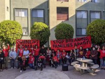 A group of tentants stand in front of a building, some with their fists in the air, with signs that say "eminent domain" and "stop breaking families apart"