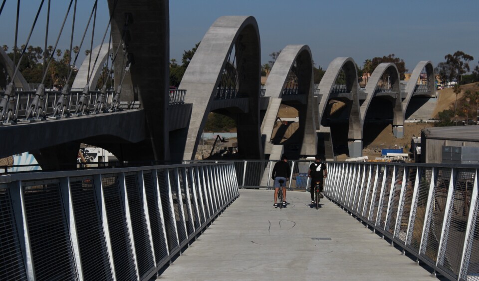 Two cyclists ride down a ramp next to the rolling concrete arches of a new bridge.