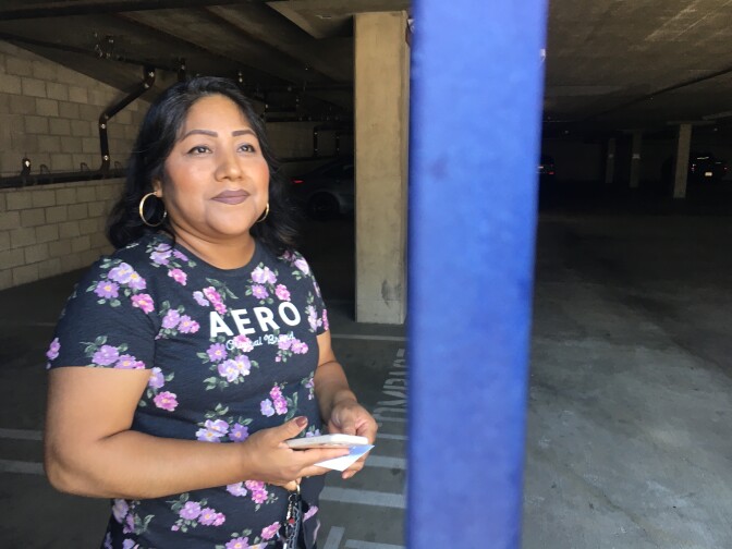 A woman with medium brown skin and long back hair, wearing gold hoop earrings and a flowered short-sleeved shirt wit the word AERO emblazoned across the front stands in an empty parking garage. A neon blue pillar is beside her.