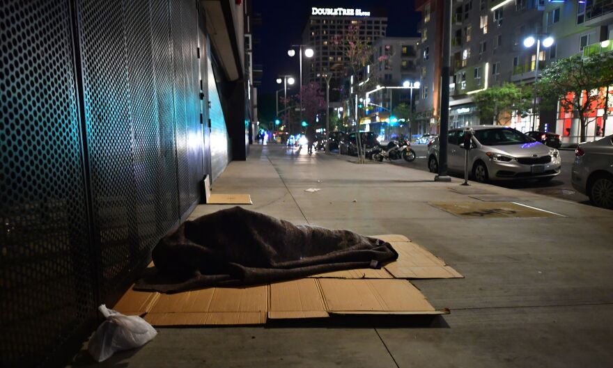 A person sleeps atop a piece of cardboard on the sidewalk covered in a blanket. The sidewalk appears to be near an active street at night in the city.