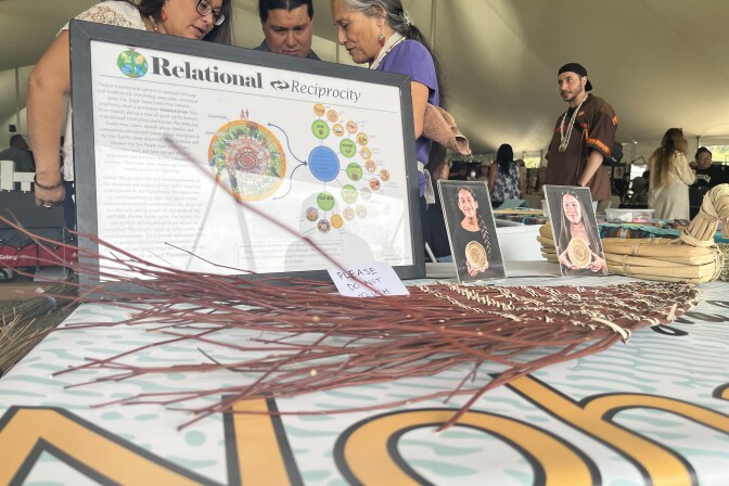 A photo on a table in the foreground reads "Relational Reciprocity" as three Native people in the background focus on an item. 