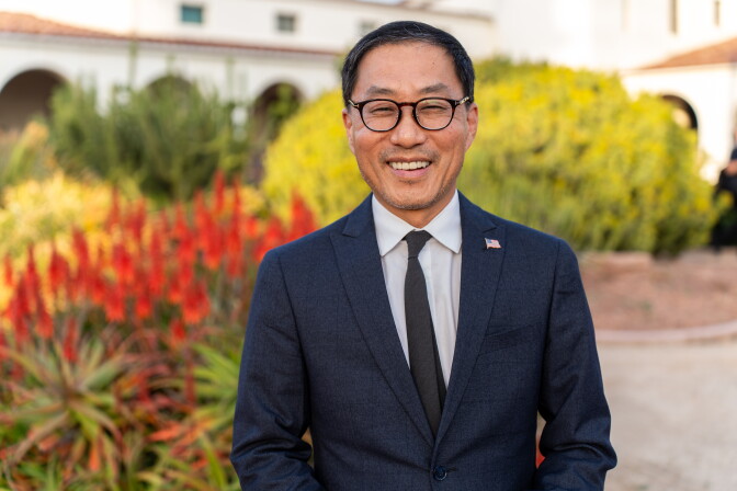 A man in glasses is wearing a dark blue suit over a white shirt and dark tie.