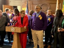 A woman in a red pantsuit with medium dark skin tone stands at a speaker podium, talking to a man with medium skin tone wearing a purple cardigan with the number 99 emblazoned above one breast.