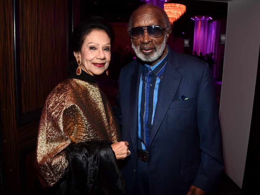 An older Black man with dark skin tone in a blue suit and sunglasses stands with his wife, who has light skin tone. They are both looking at the viewer.