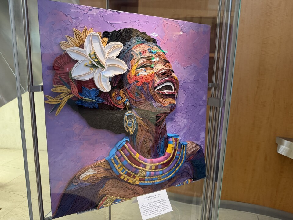 A vibrant, textured artwork displayed inside a glass case, likely in a gallery or public space. The artwork features a richly colored and highly detailed design on a square canvas with a purple background that appears to be created using a combination of techniques, including quilling or layered paper art, giving it a three-dimensional effect.