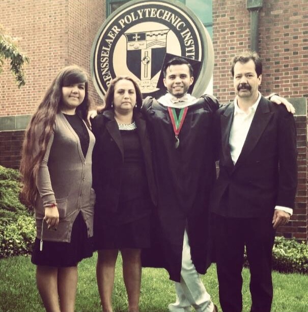 A family of four poses for a photo, including a young man in a cap and gown. They are outdoors, posing before a school seal.