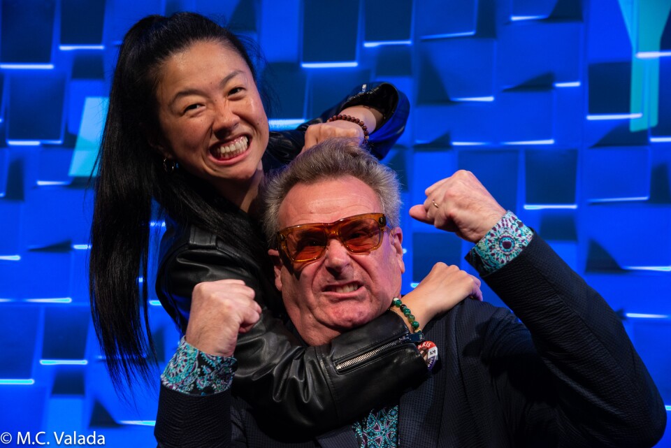 Sherry Cola, an Asian woman, is giving a friendly headlock and noogie to Greg Proops, a white man with glasses, on the LAist stage with a blue textured background