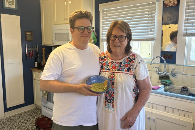 A mother and son stand next to each other inside of a kitchen that's painted blue and white. They both have white skin and are both wearing glasses. The son holds a blue bowl with a green substance inside. 