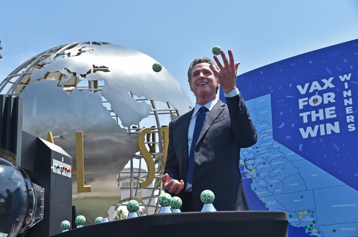 Gov. Newsom juggles numbered lottery balls in front of the Universal Studios globe and a sign touting the state's "Vax for the win" winners.